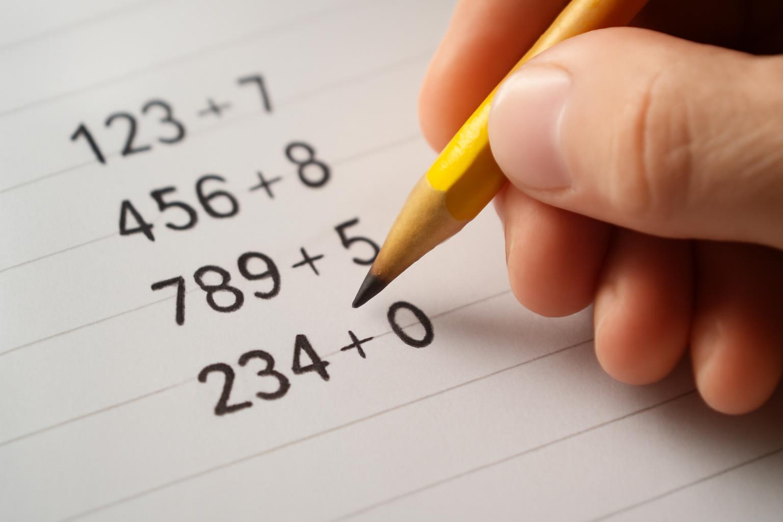 Egy kéz egy sárga ceruzával számokat ír egy papírlapra, matematikai műveletek láthatók.